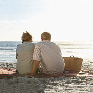 An older man and woman sitting on a picnic blanket watching the sunset on a beach