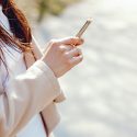 young long brown haired woman in a spring coat standing in a park texting on her mobile iphone