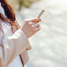 young long brown haired woman in a spring coat standing in a park texting on her mobile iphone