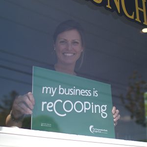 Kate, owner of Old Kings Coffee House, stands in the window of her business holding a 'my business is reCOOPing' sign