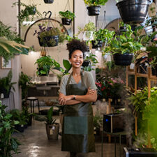 Portrait of african american woman with crossed arms wearing apron standing in botanical store. Smiling young woman in botany store standing between plants looking at camera. Happy small business owner working at flower shop standing surrounded by plants.